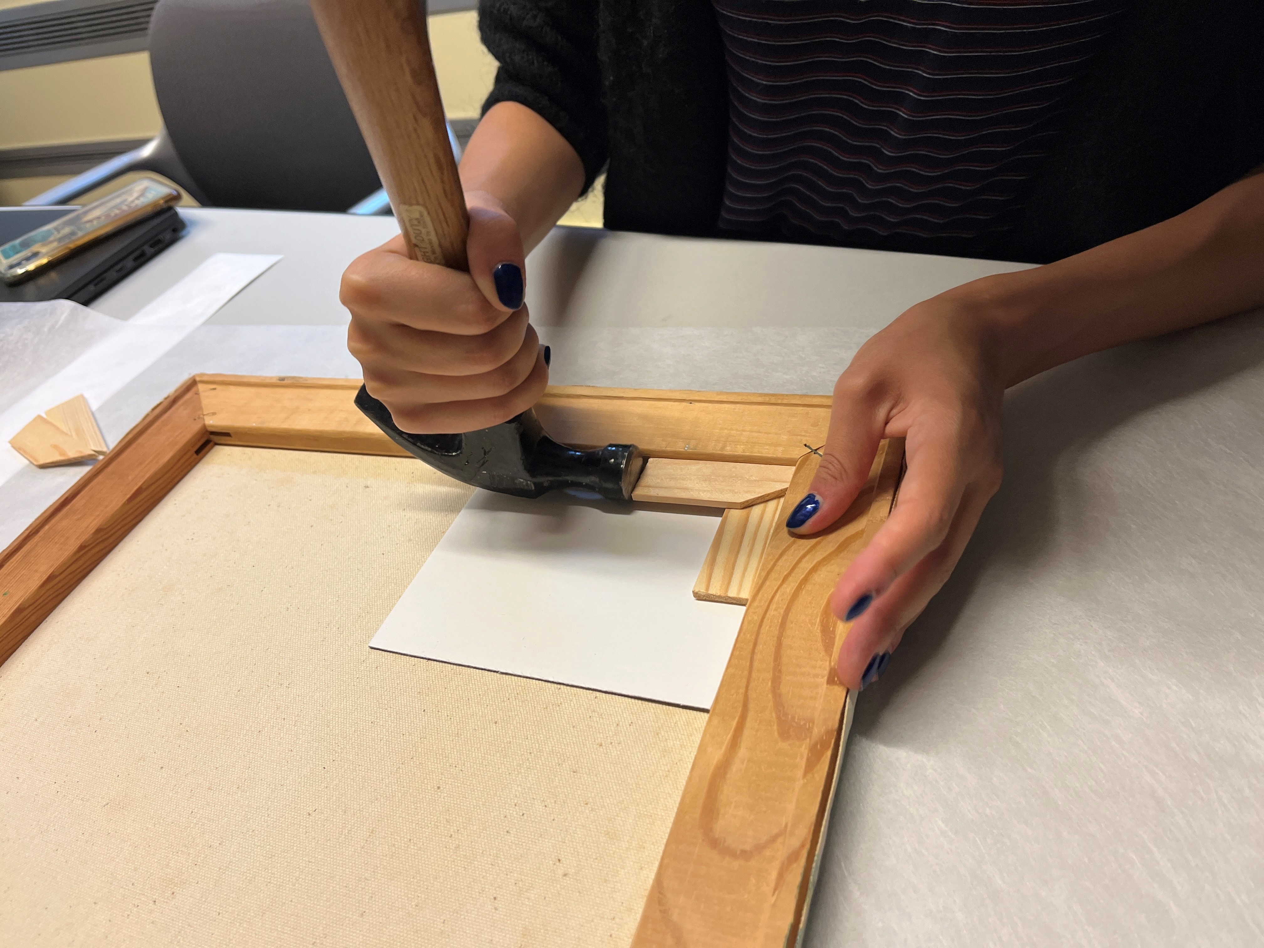 Hands holding a hammer, tapping wooden triangle keys into the corners on the back of a painting stretcher
