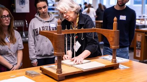A woman sewing a book on a sewing frame with students watching her progress.