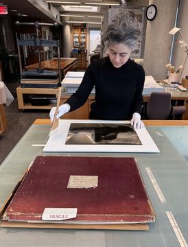 A woman wearing white gloves using a wide brush to clean a large glass photographic negative. 
