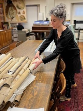 A woman taking a rolled paper item from a pile on a table in a library. 