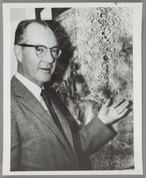 A photograph of a man wearing glasses from the 1950s standing in front of a poster of the moon.
