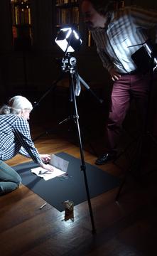 Two people, one kneeling, one standing, unfolding a document with two lights on tripods illuminating the person and document.