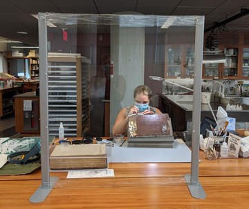 A masked woman working on a large, leather-bound volume behind a piece of plexiglass.