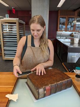 A woman in an apron measuring something on a large, leatherbound volume.