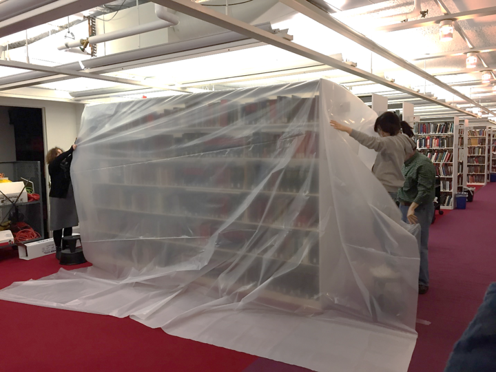 Three people draping library shelving in plastic sheeting.