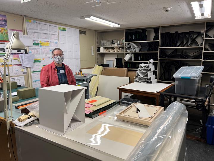 A person stands behind a desk surrounded by exhibition cradles and supplies as well as calendars and schedules.  
