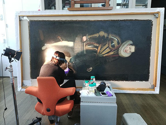 A woman sitting facing a large oil painting working on a small, lit spot on the canvas.