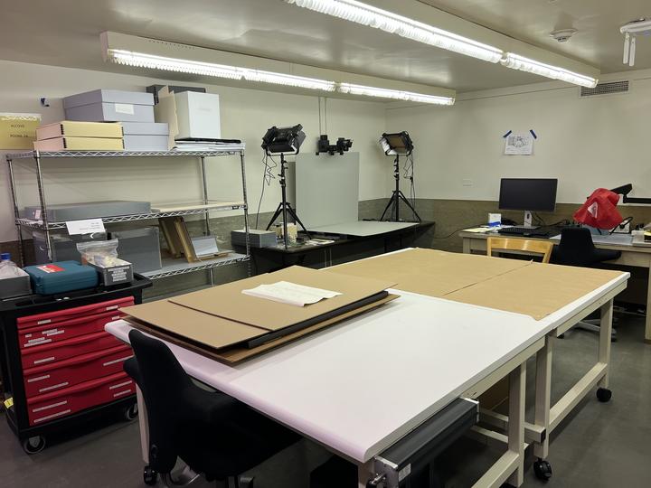 A room containing shelves, a tool cart, work tables, and a camera stand and lights.  