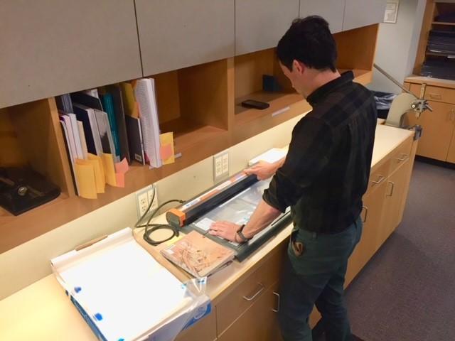 A person stands at a counter with cabinets and open shelving above using a small machine to make a book sleeve.