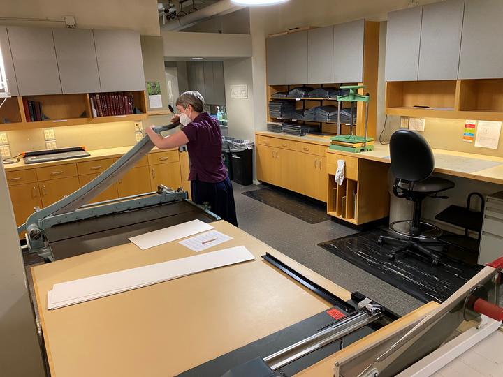 A person using a large piece of equipment for cutting board and paper. In the foreground are pieces of countertop equipment.