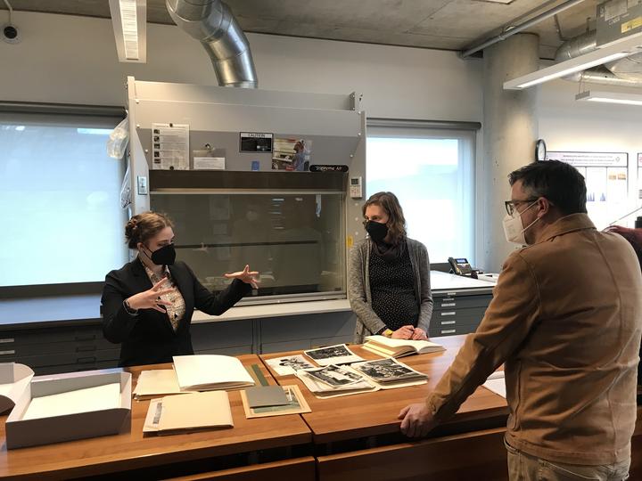 Three people standing around a table with photographs and papers laid out having a conversation.