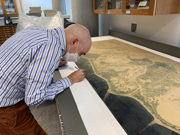 A man leaning over a large, watercolor map with some colored pencils. 