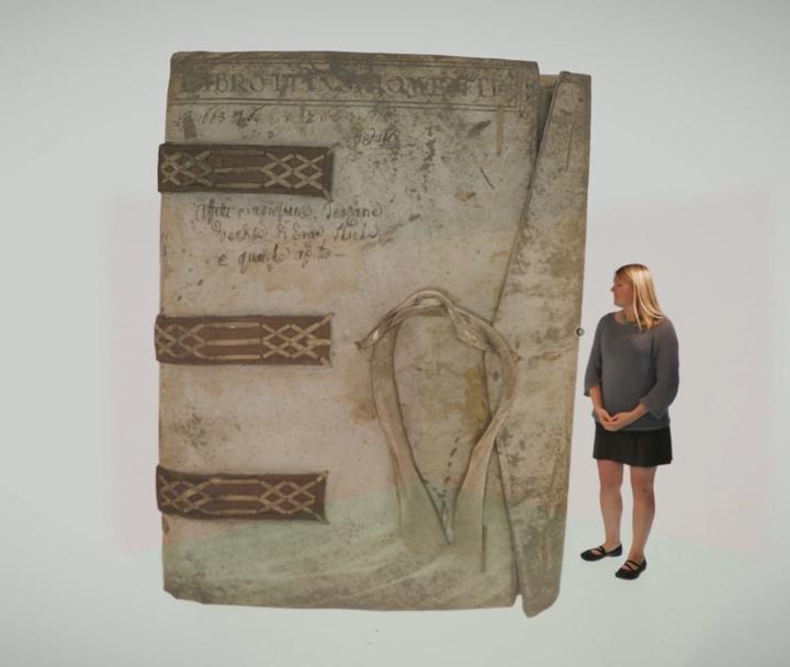 A woman standing next to an image of a large parchment-bound book.