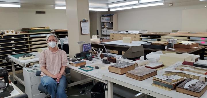 A woman sits at a long table surrounded by tools, books, and boxes.  