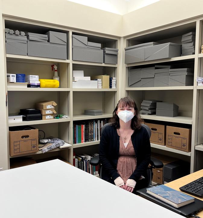 A woman sits at a desk in front of bookshelves containing tools and supplies are in the background.