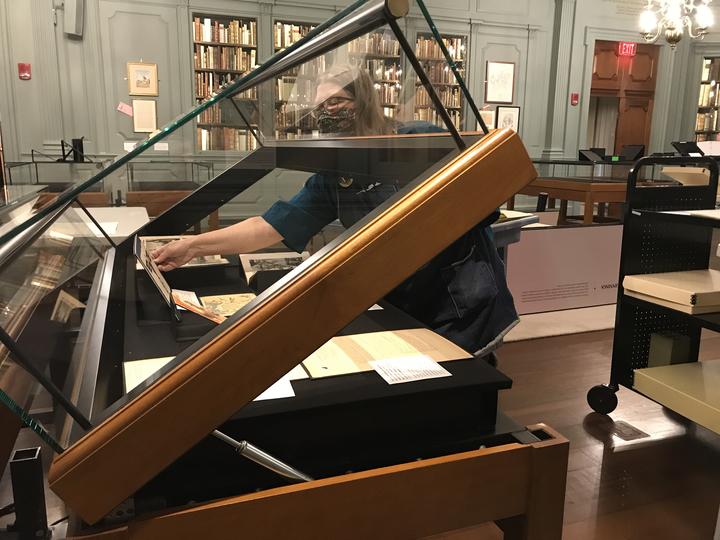 An exhibition display case lid is hinged open. A person is reaching into the case to adjust a document which is being exhibited.